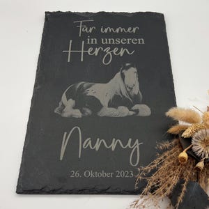 Puede incluir: Una placa rectangular de pizarra negra con una imagen grabada de un caballo tumbado. El texto dice "F&uuml;r immer in unseren Herzen", "Nanny" y "26. Oktober 2023". La placa tiene un borde de piedra natural rugoso y una superficie oscura.