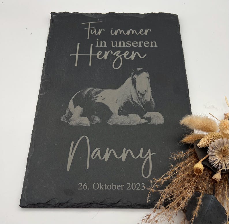 Puede incluir: Una placa rectangular de pizarra negra con una imagen grabada de un caballo tumbado. El texto dice "F&uuml;r immer in unseren Herzen", "Nanny" y "26. Oktober 2023". La placa tiene un borde de piedra natural rugoso y una superficie oscura.