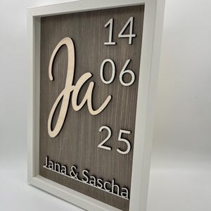 Puede incluir: Un letrero de madera enmarcado con un fondo de vetas de madera gris. El letrero presenta la palabra "Ja" en una fuente cursiva, junto con los números "14", "06" y "25". También se muestran los nombres "Jana & Sascha".