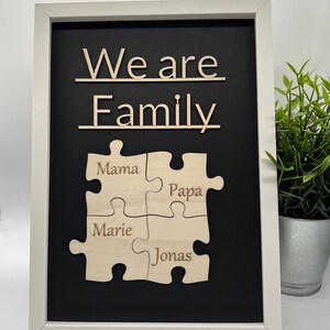 Könnte beinhalten: Ein gerahmtes Holzfamilienpuzzle mit den Worten "We are Family" oben. Darunter sind vier Puzzleteile ineinander verschlungen, die jeweils mit dem Namen eines Familienmitglieds beschriftet sind: Mama, Papa, Marie und Jonas. Der Rahmen ist weiß und der Hintergrund ist schwarz.