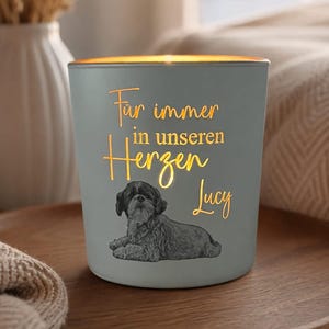 Puede incluir: Un portavelas cilíndrico de color gris claro con una luz naranja brillante que emana del interior. La parte frontal presenta el texto "Für immer in unseren Herzen Lucy" en letra cursiva sobre una ilustración detallada en blanco y negro de un perro pequeño.