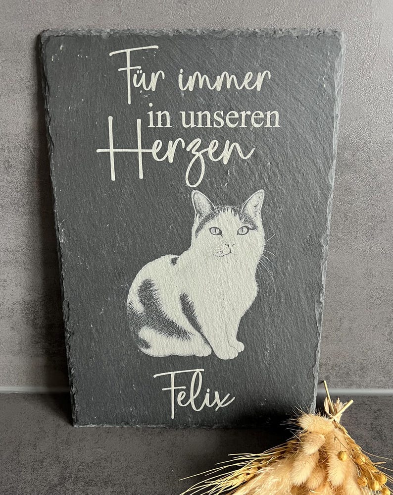 Puede incluir: Una placa rectangular de pizarra gris oscuro con una ilustraci&oacute;n grabada de un gato sentado. El texto "F&uuml;r immer in unseren Herzen" est&aacute; grabado sobre el gato, y el nombre "Felix" est&aacute; grabado debajo en una fuente blanca cursiva.