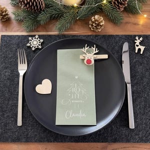 Puede incluir: Un plato llano negro mate contiene una servilleta verde salvia impresa con un árbol de Navidad blanco, el texto "Frohe Weihnachten" y el nombre "Claudia". Un clip de reno de madera con nariz roja sujeta la servilleta, flanqueado por tenedor y cuchillo.
