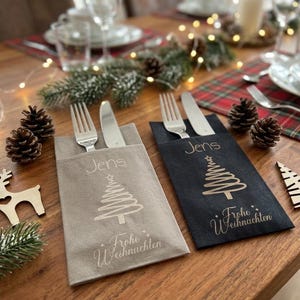 Puede incluir: Dos fundas rectangulares de tela para cubiertos, una gris claro y otra negra, cada una con un tenedor y un cuchillo de plata. Las fundas tienen impreso el nombre Jens, un gráfico de árbol de Navidad y el texto Frohe Weihnachten.