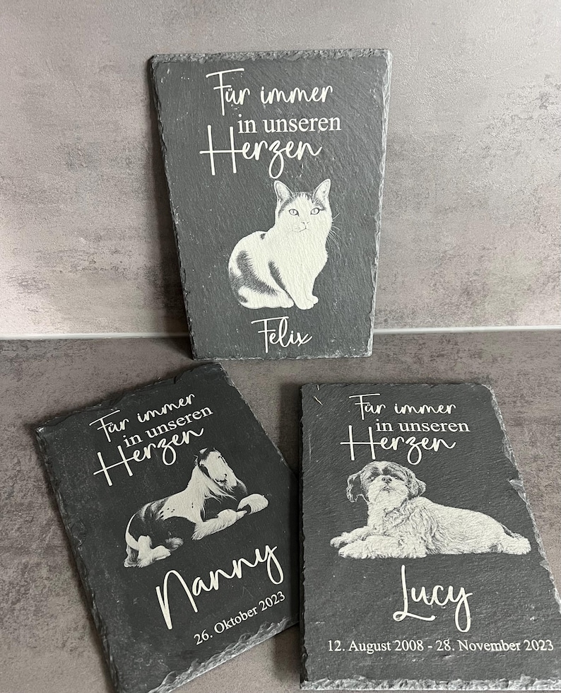 Puede incluir: Tres placas rectangulares de pizarra gris oscuro con grabados blancos de retratos de animales. Cada placa muestra la frase "F&uuml;r immer in unseren Herzen" sobre una ilustraci&oacute;n de un gato o perro y un nombre, incluyendo fechas en algunos.