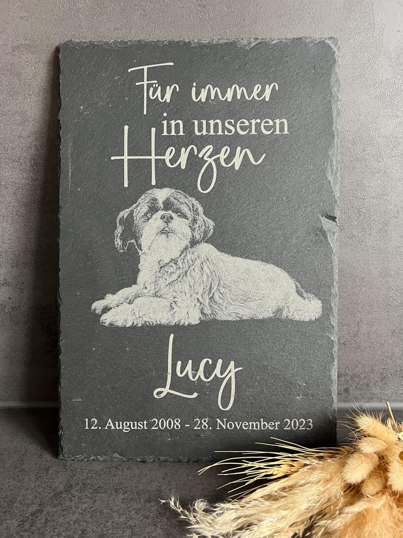 Puede incluir: Una placa rectangular de pizarra gris oscuro con la imagen grabada de un perro tumbado. El texto "F&uuml;r immer in unseren Herzen", el nombre "Lucy" y las fechas "12. August 2008 - 28. November 2023" aparecen en letras blancas.