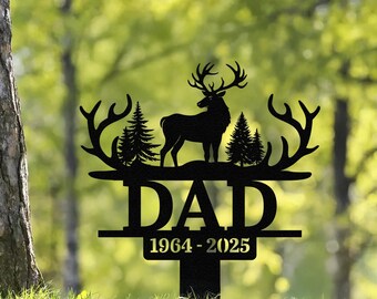 May include: Black metal yard stake featuring a deer silhouette with antlers, two pine trees, and the word "DAD" above the years "1964-2025". The stake is designed for outdoor use and is set against a blurred green background.