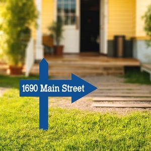 Puede incluir: Un letrero direccional azul con el texto blanco "1690 Main Street". El letrero está en un jardín con césped, con una casa amarilla y un porche al fondo.