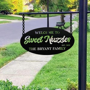 Peut inclure: Panneau ovale noir avec l'inscription "WELCOME TO Sweet Nuzzles" en écriture blanche et verte, "THE BRYANT FAMILY" et "EST. 2025". Le panneau est suspendu à un cadre en métal noir avec des chaînes.