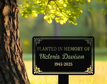 Puede incluir: Placa conmemorativa de jardín de metal negro con el texto "Planted in Memory of Victoria Davison 1945-2025". La placa tiene un borde dorado decorativo y está montada sobre una estaca negra, en una zona de césped.