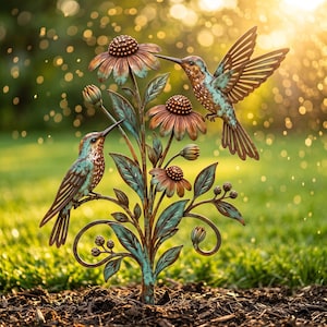 May include: A metal garden sculpture featuring two hummingbirds perched on and hovering near a flowering plant. The sculpture has a weathered copper and verdigris finish, depicting three open flowers, several buds, and detailed leaves on stems.