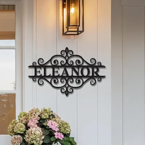 Può includere: Cartello in metallo nero con il nome "ELEANOR" in un carattere decorativo. Il cartello è montato su un muro bianco, con un'apparecchiatura di illuminazione sopra e un mazzo di fiori sotto. Il cartello ha un design ornato a volute.