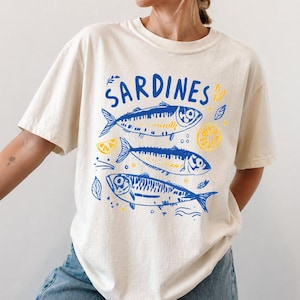 Woman wearing an ivory Comfort Colors t-shirt featuring a blue vintage sardines illustration with lemons in a Mediterranean coastal style.
