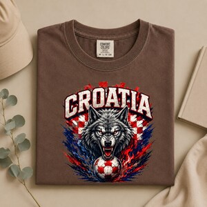 May include: A brown short-sleeve t-shirt with a graphic print featuring a snarling wolf head above a soccer ball. The design includes the text CROATIA in white block letters over a red and white checkered pattern with blue and red flame accents.