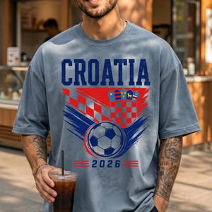 May include: A slate blue t-shirt featuring the word CROATIA in bold blue letters above a red and white checkered crest, a soccer ball graphic, and the year 2026 in blue text with red horizontal lines on either side of the date.