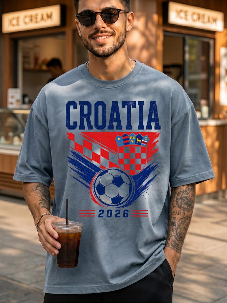 May include: A slate blue t-shirt featuring the word CROATIA in bold blue letters above a red and white checkered crest, a soccer ball graphic, and the year 2026 in blue text with red horizontal lines on either side of the date.