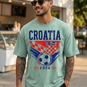 May include: A light green short-sleeve t-shirt featuring the text CROATIA in blue, a red and white checkered pattern, a shield emblem, a soccer ball graphic, and the year 2026 printed in blue with red horizontal lines on the front.