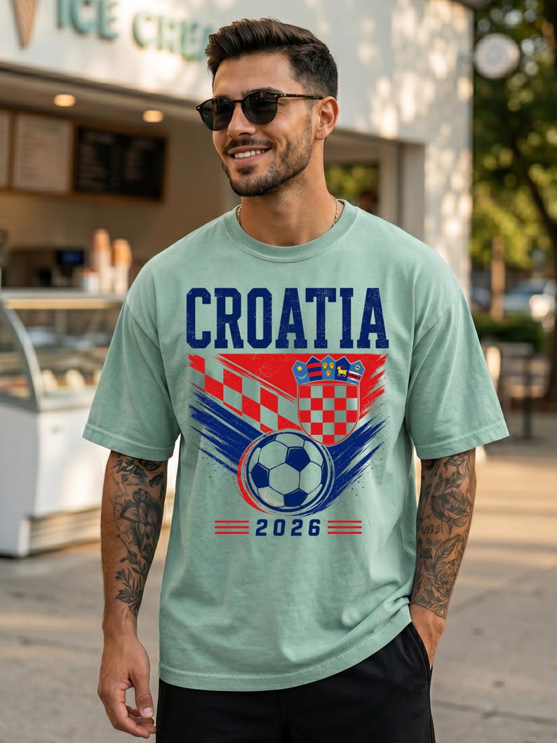May include: A light green short-sleeve t-shirt featuring the text CROATIA in blue, a red and white checkered pattern, a shield emblem, a soccer ball graphic, and the year 2026 printed in blue with red horizontal lines on the front.