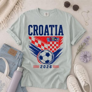 May include: A light gray short-sleeve t-shirt featuring the text CROATIA in blue, a red and white checkered pattern, a soccer ball graphic, the Croatian coat of arms, and the year 2026 printed in blue and red on the front of the garment.