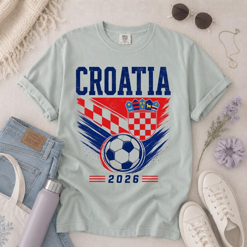 May include: A light gray short-sleeve t-shirt featuring the text CROATIA in blue, a red and white checkered pattern, a soccer ball graphic, the Croatian coat of arms, and the year 2026 printed in blue and red on the front of the garment.