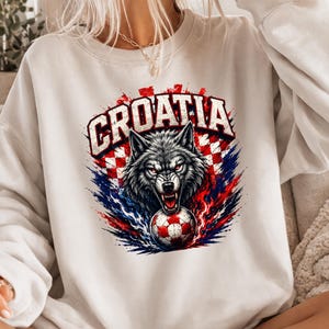 May include: A white crewneck sweatshirt featuring a graphic print of a snarling wolf head above a soccer ball. The design includes the text CROATIA in bold letters, set against a red and white checkered pattern with blue and red paint splatter effects.