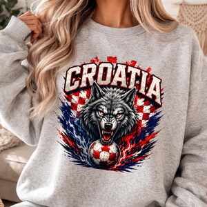May include: A light gray crewneck sweatshirt featuring a graphic print of a snarling wolf head above a soccer ball. The design includes the text CROATIA in bold white letters, set against a red and white checkered pattern with blue accents.