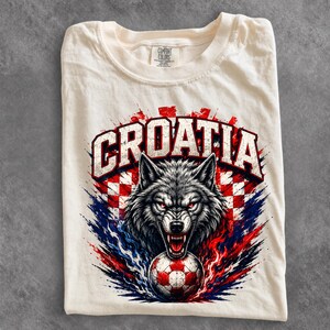 May include: A cream-colored t-shirt featuring a graphic print of a snarling wolf head above a soccer ball. The text CROATIA is arched above the wolf in bold white letters with a red and white checkered background and blue and red paint splatters.