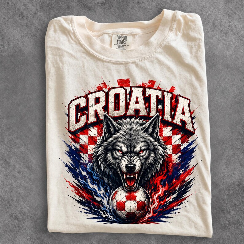 May include: A cream-colored t-shirt featuring a graphic print of a snarling wolf head above a soccer ball. The text CROATIA is arched above the wolf in bold white letters with a red and white checkered background and blue and red paint splatters.