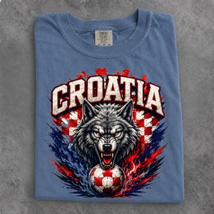May include: A blue crew neck t-shirt featuring a graphic of a snarling wolf head above a soccer ball. The word CROATIA is printed in large white letters above the wolf. The background design includes red and white checkered patterns and blue flames.