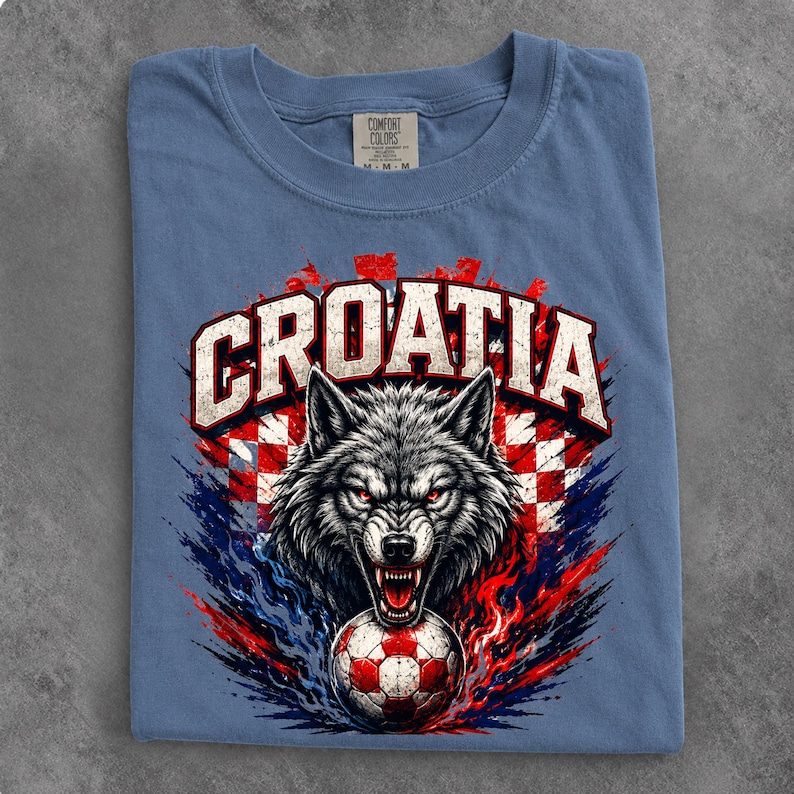 May include: A blue crew neck t-shirt featuring a graphic of a snarling wolf head above a soccer ball. The word CROATIA is printed in large white letters above the wolf. The background design includes red and white checkered patterns and blue flames.