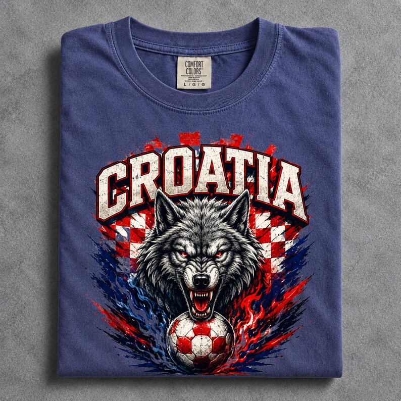 May include: A blue t-shirt featuring a graphic of a snarling wolf head above a soccer ball. The word CROATIA is printed in large white letters above the wolf. The design includes red and white checkered patterns and blue and red paint splatters.