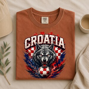 May include: A rust-colored t-shirt featuring a graphic of a snarling wolf head above a soccer ball. The word CROATIA is printed in large white letters above the wolf. The background of the graphic includes a red and white checkered pattern.