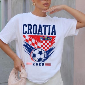 May include: A white short-sleeve t-shirt featuring the text CROATIA in blue, a red and white checkered pattern, the Croatian coat of arms, a soccer ball graphic, and the year 2026 printed in blue with red horizontal lines on each side.