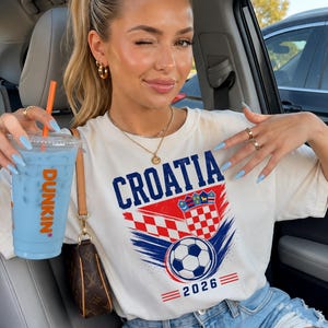 May include: A cream-colored short-sleeve t-shirt featuring the text CROATIA in blue, a red and white checkered crest, a soccer ball graphic, and the year 2026 in blue, accompanied by a clear plastic cup containing a light blue drink and an orange straw.