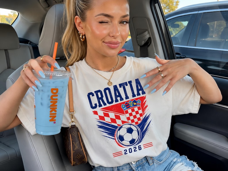 May include: A cream-colored short-sleeve t-shirt featuring the text CROATIA in blue, a red and white checkered crest, a soccer ball graphic, and the year 2026 in blue, accompanied by a clear plastic cup containing a light blue drink and an orange straw.