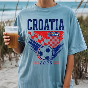 May include: A light blue t-shirt featuring the text CROATIA in dark blue letters above a graphic of the Croatian coat of arms, a soccer ball, and red and blue brushstroke accents. The number 2026 is printed in blue at the bottom of the design.