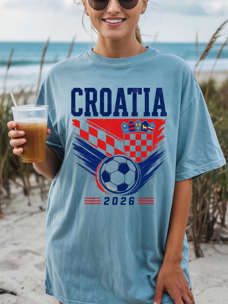 May include: A light blue t-shirt featuring the text CROATIA in dark blue letters above a graphic of the Croatian coat of arms, a soccer ball, and red and blue brushstroke accents. The number 2026 is printed in blue at the bottom of the design.