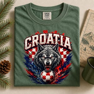 May include: An olive green t-shirt featuring a graphic of a snarling wolf head above a soccer ball. The text CROATIA is printed in white block letters above the wolf. The design includes a red and white checkered pattern and blue accents.