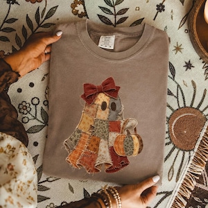 May include: A taupe-colored crewneck t-shirt featuring a central graphic of a ghost made of various patchwork fabric patterns in autumn colors. The ghost wears a dark red bow and carries a small pumpkin. The shirt has a Comfort Colors label.
