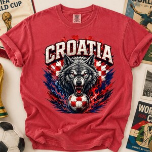 May include: A red short-sleeve t-shirt featuring a graphic of a snarling wolf head above a soccer ball. The text CROATIA is printed in white above the wolf. The design includes red and white checkered patterns and blue flame accents.