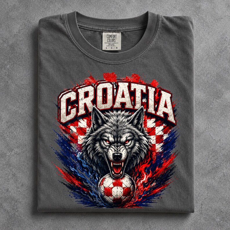 May include: A charcoal gray t-shirt featuring a graphic of a snarling wolf with red eyes, positioned above a soccer ball. The design includes the text CROATIA in white block letters, set against a red and white checkered pattern and blue accents.