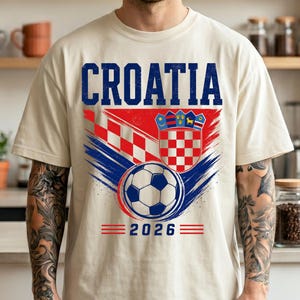May include: A cream-colored short-sleeve t-shirt featuring the text CROATIA in blue, a red and white checkered pattern, the Croatian coat of arms, a soccer ball graphic, and the year 2026 printed in blue with red horizontal lines on the sides.