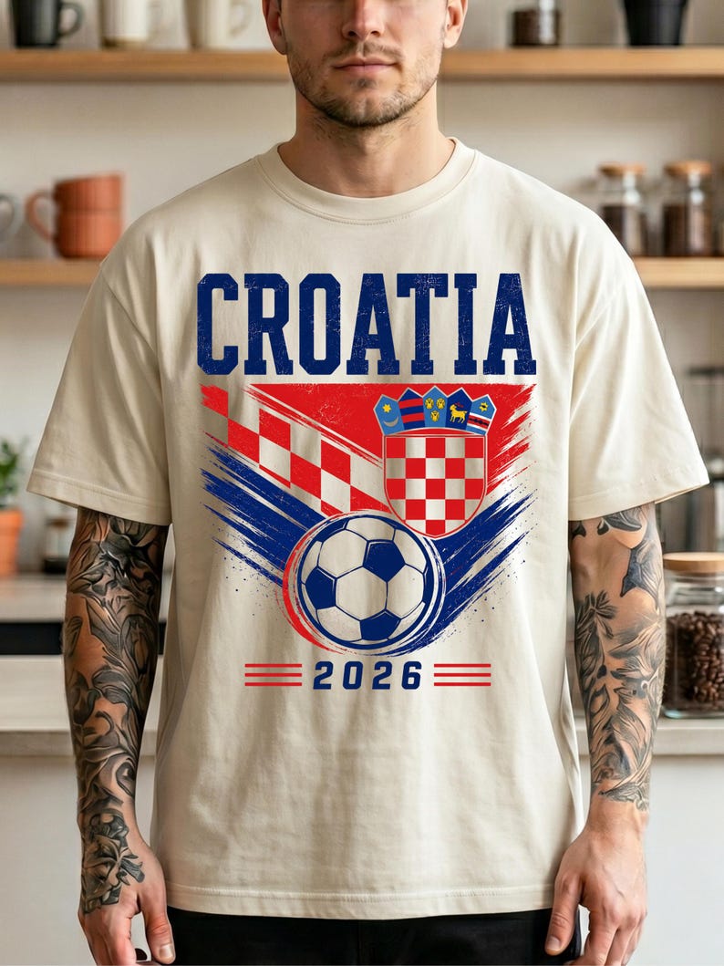 May include: A cream-colored short-sleeve t-shirt featuring the text CROATIA in blue, a red and white checkered pattern, the Croatian coat of arms, a soccer ball graphic, and the year 2026 printed in blue with red horizontal lines on the sides.