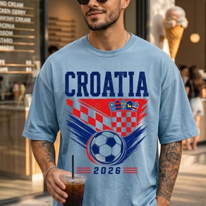 May include: A light blue t-shirt featuring the word CROATIA in dark blue letters above a graphic of the Croatian coat of arms, a soccer ball, and the year 2026. The design includes red and blue brushstroke accents and a red checkered pattern.