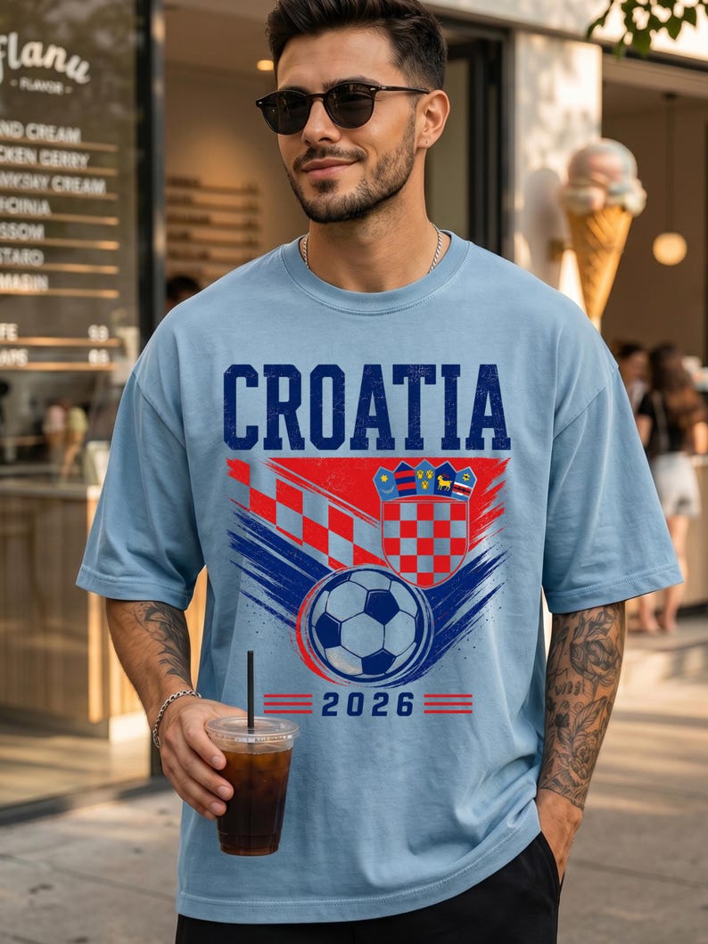 May include: A light blue t-shirt featuring the word CROATIA in dark blue letters above a graphic of the Croatian coat of arms, a soccer ball, and the year 2026. The design includes red and blue brushstroke accents and a red checkered pattern.