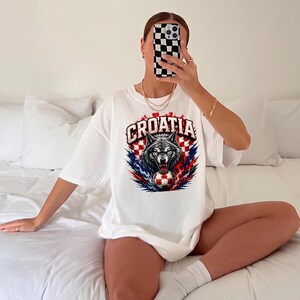 May include: A white short-sleeved t-shirt featuring a graphic print of a wolf head with a soccer ball in its mouth. The design includes the text CROATIA in bold letters above the wolf, set against a red and white checkered pattern background.