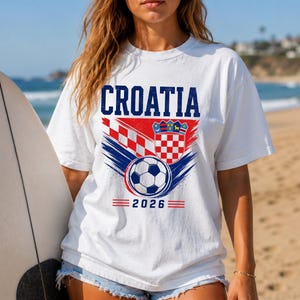 May include: A white short-sleeve t-shirt featuring the text CROATIA in blue, a red and white checkered pattern, a shield emblem, and a soccer ball graphic above the year 2026 in blue text with red horizontal lines on either side of the date.