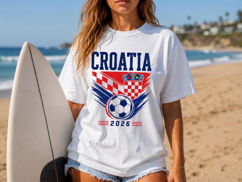 May include: A white short-sleeve t-shirt featuring the text CROATIA in blue, a red and white checkered pattern, a shield emblem, and a soccer ball graphic above the year 2026 in blue text with red horizontal lines on either side of the date.
