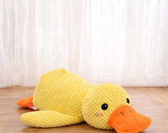 Puede incluir: Un juguete de peluche de pato amarillo con pico y patas naranjas, tumbado en un suelo de madera. El pato tiene ojos negros y mejillas rosas. El juguete está hecho de una tela texturizada.