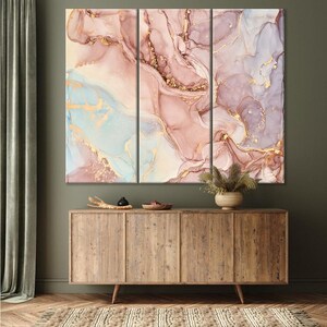 May include: A triptych of abstract art panels featuring a fluid design in shades of pink, lavender, and light blue, accented with gold veins. The artwork is displayed above a wooden cabinet.